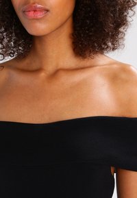 Haut noir à épaules dénudées en tissu lisse et texturé. Il présente une large bande et une silhouette ajustée, mettant en valeur la clavicule.