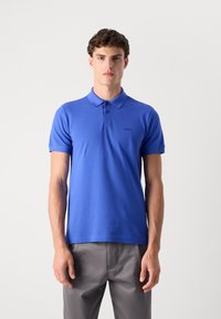 Camisola polo azul feita de algodão, com gola, três botões e mangas curtas. Detalhe sutil do logótipo no lado esquerdo do peito. Combinada com calças cinzas.