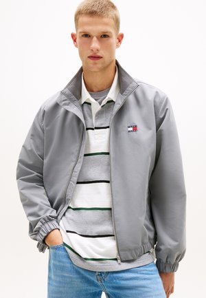 Joven con chaqueta gris clara con cremallera sobre una camisa polo a rayas y jeans azules, de pie con una mano en el bolsillo.