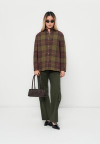 Rutig oversized skjorta i grönt och vinrött, olivgröna vida byxor, svarta solglasögon och en mörkbrun handväska med korta handtag.