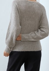 Personne portant un pull en maille beige avec des motifs subtils et de petites perles sur la manche, associé à un pantalon noir, la main cachée derrière le dos.