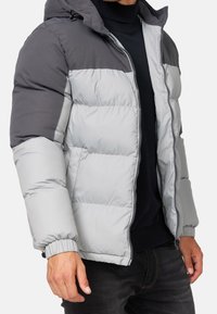 Veste matelassée grise et gris clair avec capuche, présentant une texture matelassée, des poignets élastiques et une fermeture éclair à l'avant.