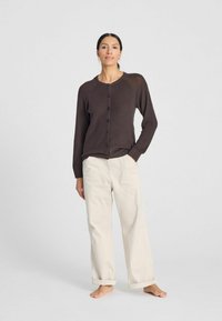 Brauner, knopfbarer Cardigan mit langen Ärmeln, aus leichtem Stoff gefertigt. Kombiniert mit locker sitzenden, cremefarbenen Hosen, barfuß getragen.