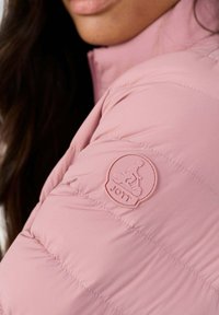 Chaqueta acolchada rosa con textura suave, diseño acolchado y un distintivo parche de logo redondo en la manga.