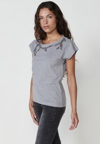 Grå kortärmad T-shirt i mjukt material, med dekorativa utsmyckningar längs halsringningen och en klassisk passform.