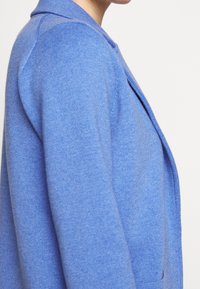 Blåstickad cardigan med texturerad tyg, strukturerad krage och figursydd silhuett, med synliga sid sömmar och minimal detaljer.