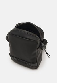 Černá taška s měkkou texturou a čtvercovým tvarem, se zipovým uzávěrem. Na přední straně má logo Tommy Hilfiger a látkový popruh pro nošení.