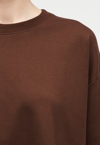 Κοντινό πλάνο ενός ατόμου που φοράει ένα απλό καφέ φούτερ με στρογγυλή λαιμόκοψη και εμφανή ραφή γύρω από το λαιμό και τον ώμο.