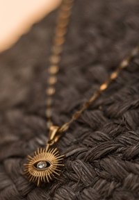 Gold necklace featuring a sunburst pendant with a central eye design and a small clear gemstone, set against a textured dark background.