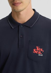 Camisa polo azul marino con cuello texturizado. Presenta un texto gráfico rojo "Sí, Mar." y "LIU·JO" en el bolsillo. Cuello de dos botones con botones de contraste.