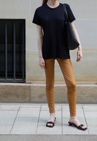 T-shirt noir à manches courtes associé à un pantalon ajusté beige et des sandales à glisser noires, sur fond de mur en pierre claire et de pavé.