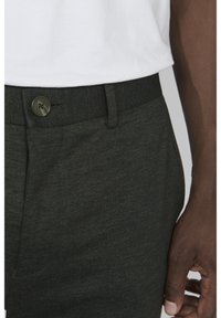 Pantalones verde oscuro con una tela texturada, que cuentan con un cierre de botón y trabillas, parcialmente cubiertos por una camisa blanca.