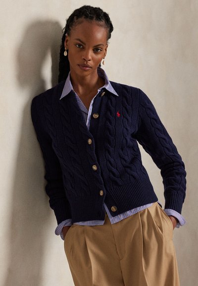 Mujer con el pelo trenzado que lleva un cárdigan azul marino de punto trenzado sobre una camisa azul a rayas y pantalones beige, de pie frente a una pared lisa.