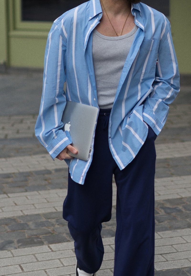 Camisa de botones a rayas azul claro y blanca, camiseta gris sin mangas debajo, pantalones sueltos azul marino, sosteniendo una computadora portátil plateada, caminando sobre una superficie pavimentada.