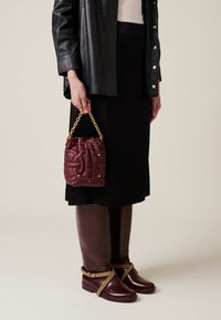 Maroon leather bucket bag with drawstring closure, gold chain handle, and stud accents. Paired with a black skirt and tall brown boots.
