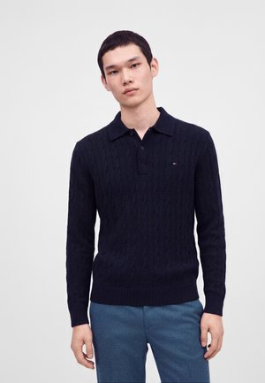 Jeune homme portant un pull polo à manches longues en maille torsadée bleu marine foncé et un pantalon bleu, debout devant un fond clair uni.