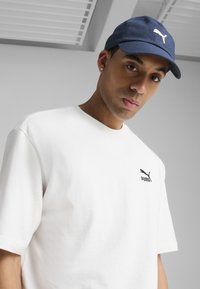 Hvid bomulds T-shirt med korte ærmer, der har et sort Puma-logo på brystet; kombineret med en blå kasket, der viser et hvidt Puma-emblem.