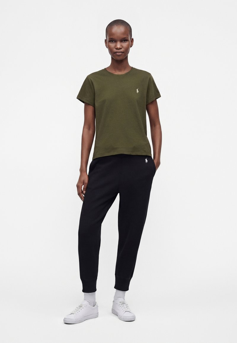 Femme debout portant un t-shirt manches courtes vert olive, un pantalon de jogging noir, des chaussettes blanches et des baskets blanches sur fond blanc.