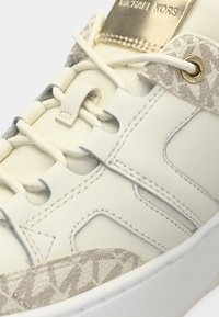 Beli usnjeni športni copat z reliefnim kremnim poudarkom, ravnimi vezalkami in zlatim logotipom. Podrobni šivi in vzorčasti boki.