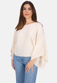 Helle Creme, strukturierter Oberteil mit weiten Ärmeln und Spitzenbesatz. Kombiniert mit blauen Jeans, präsentiert einen entspannten, lässigen Stil.