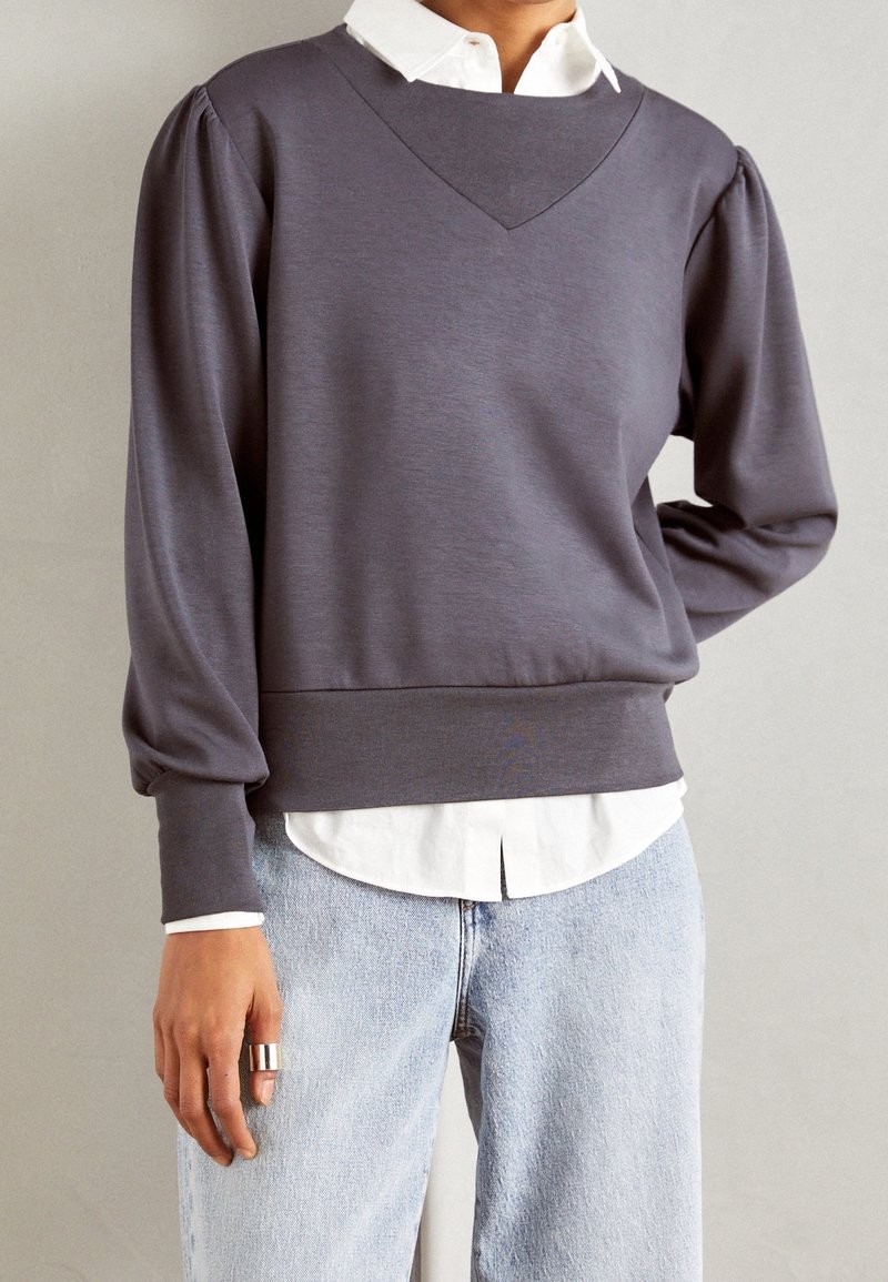 Persoon in een grijze lange mouw top over een witte shirt, gestoken in lichtblauwe jeans, met een zilveren ring om hun linkerhand.