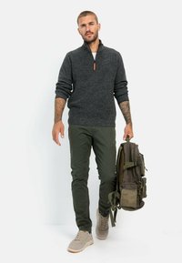 Ciemnoszary sweter na zamek, oliwkowe spodnie, beżowe sneakersy, trzymający ciemnozielony plecak z wieloma kieszeniami, stojący na białym tle.