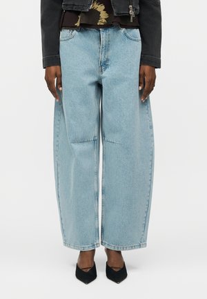 Άτομο που φοράει ανοιχτό μπλε τζιν με φαρδιές παντελόνες, μαύρα τακούνια με μυτερή μύτη και μαύρο σακάκι πάνω από ένα φλοράλ τοπ, στέκεται μπροστά σε μια απλή φόντο.