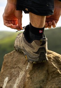 Zapato de senderismo marrón con una parte superior texturizada, calcetín de patrón negro y rojo, y una superficie rocosa; persona atándose los cordones, al aire libre con vegetación.