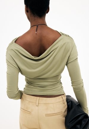 Osoba v svetlo olivovom topánku s dlhými rukávmi a zdobeným chrbtom, oblečená v béžových nohaviciach, drží čiernu koženú bundu v pravej ruke.