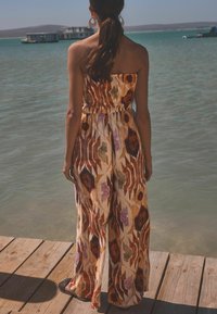 Bystövlad jumpsuit med ett flerfärgat blommönster i jordnära toner, samlad midja och vida ben, bärs med flata sandaler på en träbrygga.