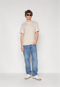 Stripet t-skjorte med korte ermer i beige og hvitt, kombinert med blå jeans. Merkbare detaljer inkluderer en liten logo på brystet og solbriller.