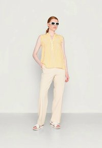 Blusa sem mangas amarela e branca com riscas, com colarinho, combinada com calças largas em bege claro e sandálias brancas, acessorizada com óculos de sol brancos.