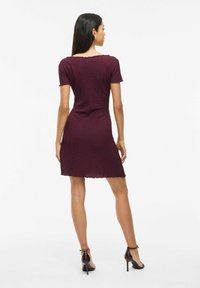 Vestido de manga corta en burdeos con tejido texturizado, dobladillo en forma de pico y silueta ajustada, combinado con zapatos de tacón alto con tiras.