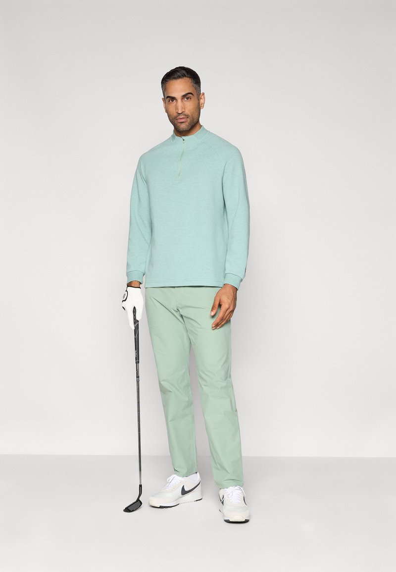 Mintgroen shirt met lange mouwen en een halve ritssluiting bij de kraag, gecombineerd met lichtgroene broek. De persoon houdt een golfclub vast en draagt witte sneakers.