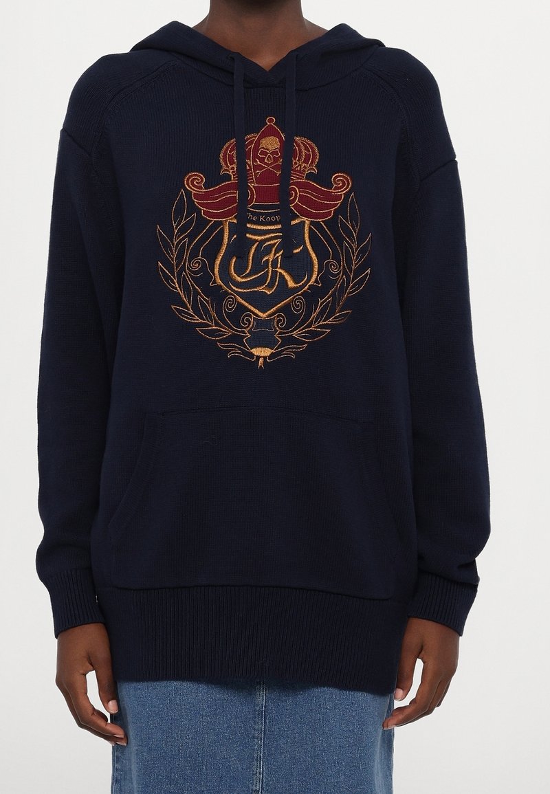 Sweat à capuche bleu marine avec un tricot texturé, arborant une crête brodée en or et rouge avec un design de couronne et de laurier, et une poche avant.