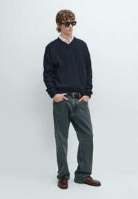 Maglione a coste blu navy, camicia bianca con colletto, jeans grigi larghi, scarpe in pelle marrone e occhiali da sole neri indossati da una persona su uno sfondo chiaro.