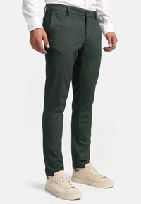 Grüne Hosen im Fischgrätenmuster mit tailliertem Schnitt und Fronttaschen, kombiniert mit beigefarbenen Sneakers. Glatte Textur, mittelhoher Bund.