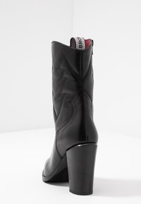 Bottine en cuir noir avec un talon haut empilé, piqûres décoratives en zigzag et une languette en tissu en haut.