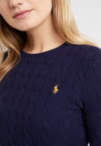 Gros plan d’une femme portant un pull en maille torsadée bleu marine avec un petit logo brodé jaune de joueur de polo sur la poitrine.