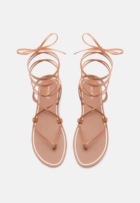 Tan kleurige sandaaltjes met een ingewikkeld ontwerp, voorzien van dunne, golvende banden en een platte zool, benadrukt door een contrasterende witte rand.