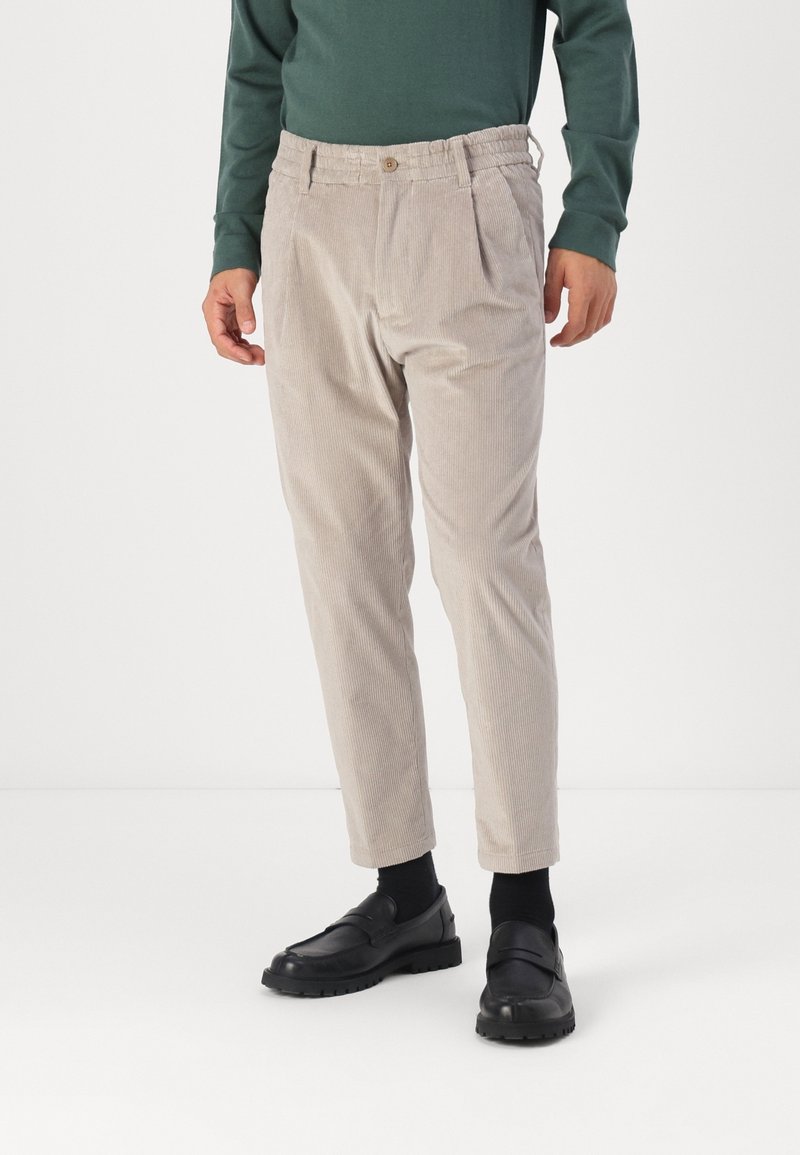 Pantalon en velours côtelé beige avec taille élastique et bouton, porté avec des chaussettes noires, des mocassins noirs et un haut à manches longues vert foncé.