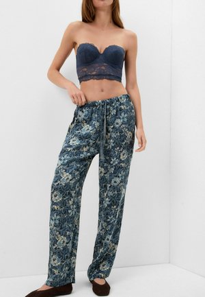Femme portant un top bustier en dentelle bleue et un pantalon ample à motif floral avec des chaussures marron devant un mur blanc.
