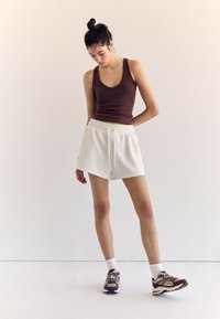 Pantaloni corti bianchi sportivi con cordoncino, abbinati a una canotta marrone e scarpe da ginnastica con motivo. Texture liscia e design casual ben evidenti.