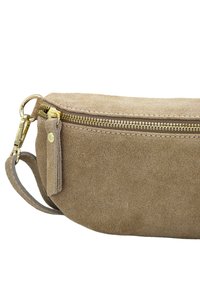 Le sac en suède brun présente une fermeture éclair et des éléments en or, avec une forme arrondie et une texture douce, complétée par une petite touche en cuir.