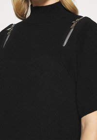 Pull noir à col montant avec manches courtes, doté de fermetures éclair argentées sur les épaules. Texture douce, coupe décontractée et design épuré.