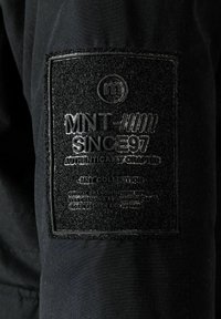 Un patch textile noir avec un branding en relief "MNT" et "SINCE 97", accompagné d'accents texturés, cousu sur une surface en tissu sombre.