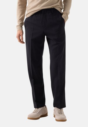 Mann trägt schwarze gerade geschnittene Hosen mit elastischem Bund und beige Sneakers, steht mit einer Hand in der Tasche.