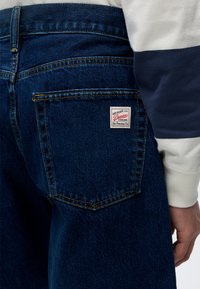 Gros plan de la poche arrière d'un jean denim bleu foncé avec une étiquette blanche rectangulaire GAP Denim Premier et partie d'une manche blanche à rayures bleu marine.