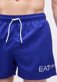 Blå badeshorts med hvid snor og EA7 Emporio Armani-logo, båret af en person, der viser talje og overben.