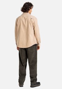 Camicia beige a maniche lunghe, vestibilità comoda, polsini con bottoni, dettaglio sullo scollo posteriore. Pantaloni in corduroy verde scuro, gamba ampia, due tasche posteriori, abbinati a scarpe nere.